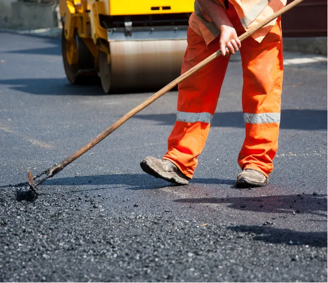 asphalt-maintenance-1 Worker in orange clothing spreads asphalt with a tool as a road roller compacts the surface behind them.