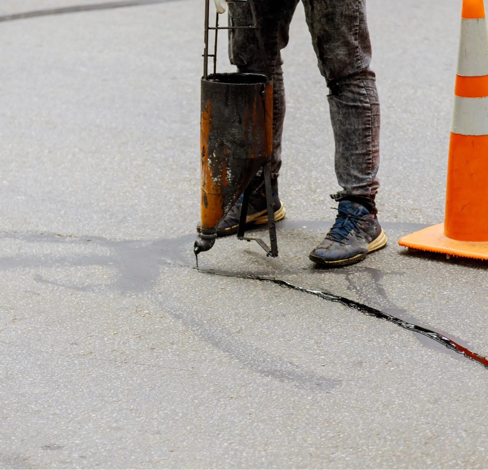 Worker sealing a road crack with equipment near an orange traffic cone on an asphalt surface.