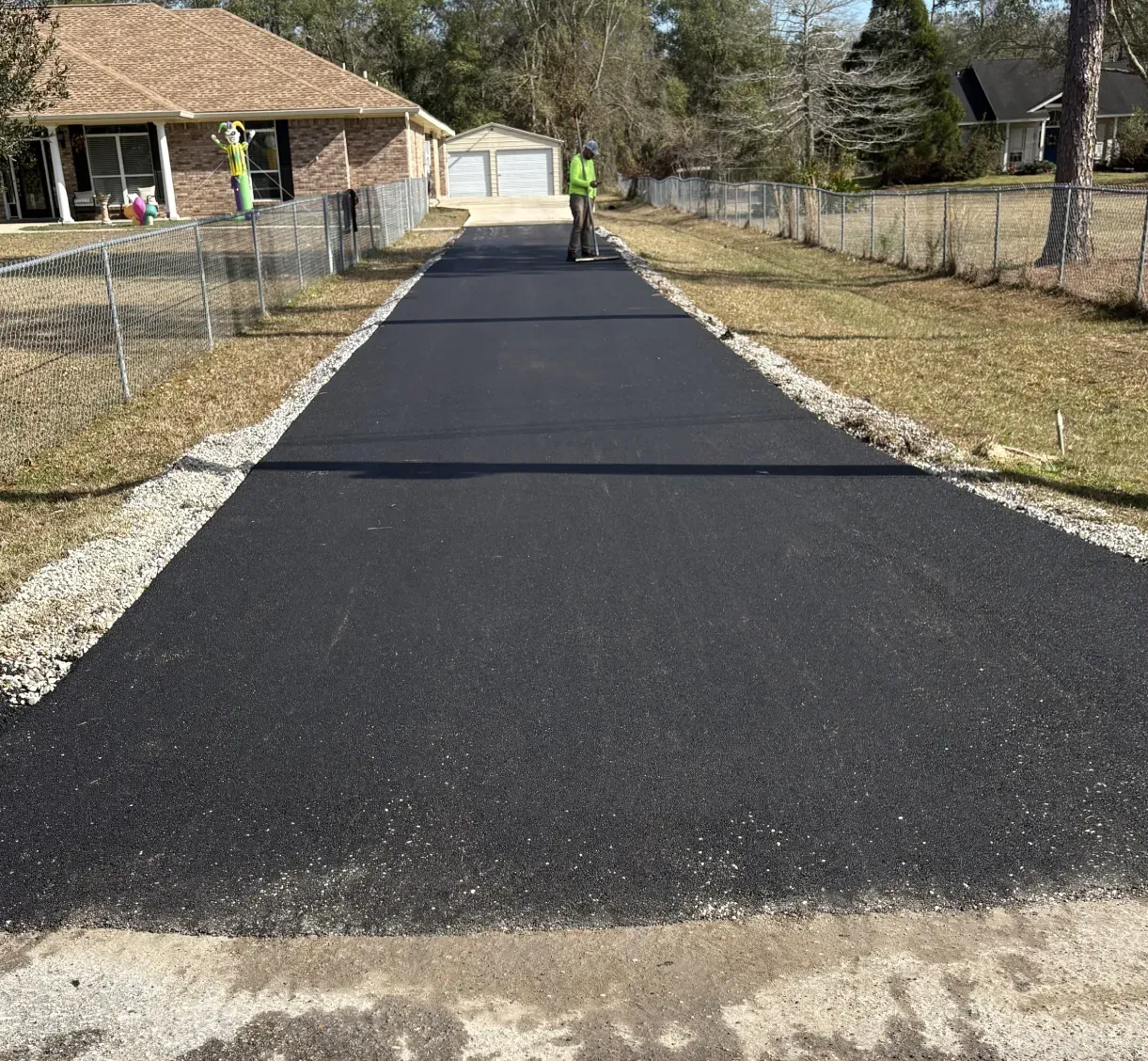 driveway-2 Freshly paved asphalt driveway with two workers standing on it, between fenced grassy yards and houses.