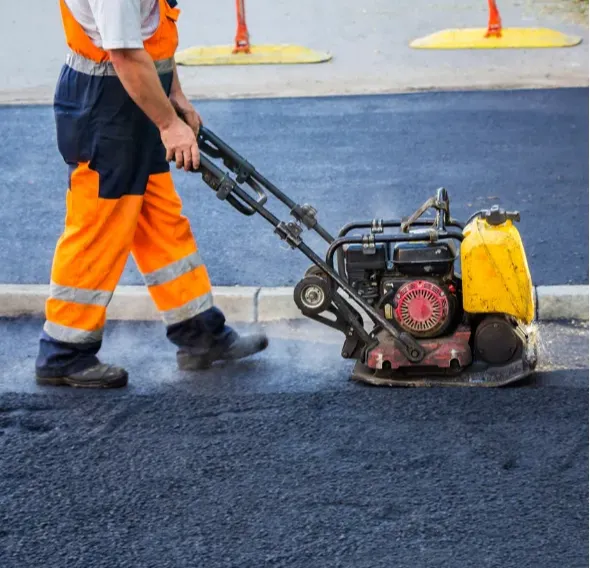maintenance-2 A worker in orange safety gear operates a compactor on freshly laid asphalt pavement.