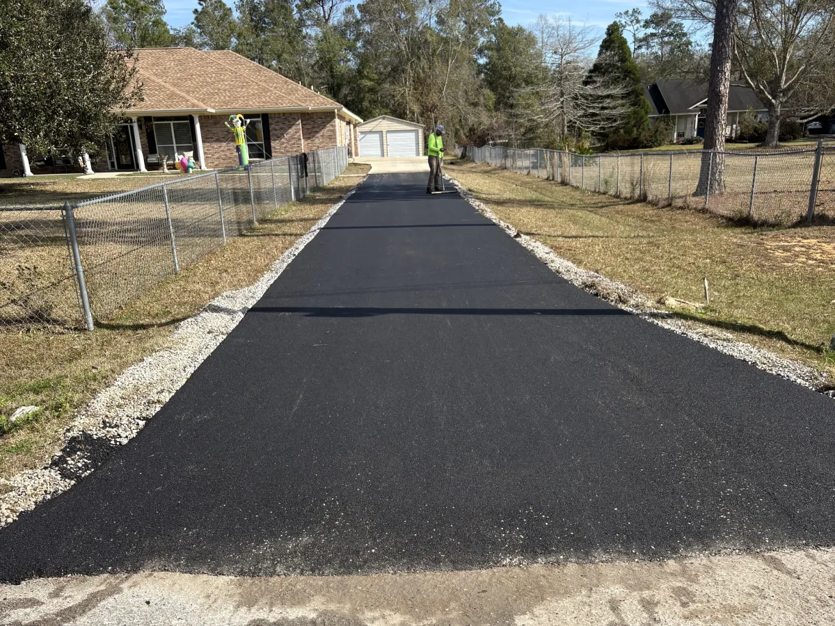 Newly paved asphalt driveway with workers in safety vests, leading to a house and garage on a sunny day.