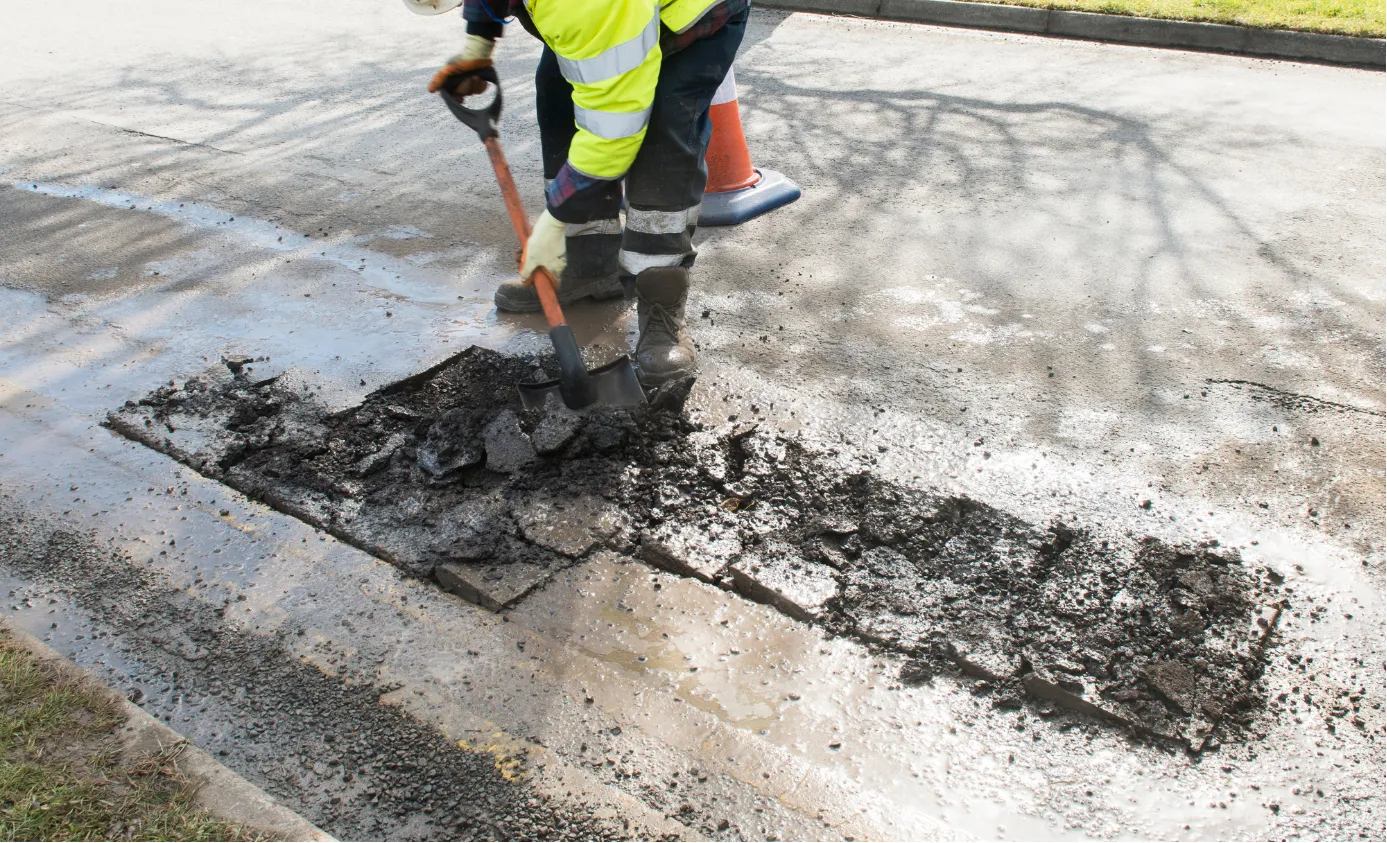 Worker in safety gear using a shovel to dig up asphalt on a road, with traffic cones nearby.