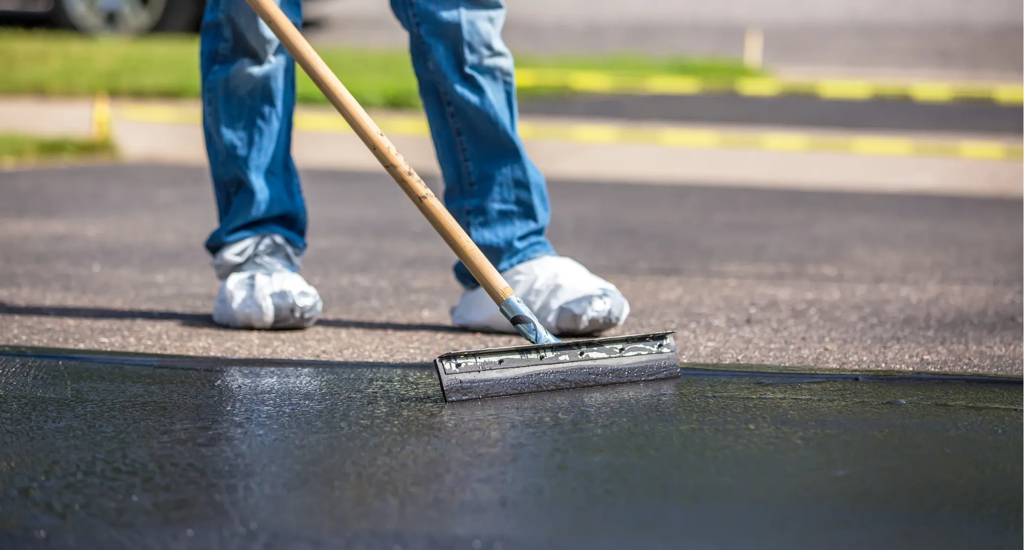 Person in jeans applying a black sealant to a driveway with a long-handled squeegee tool.