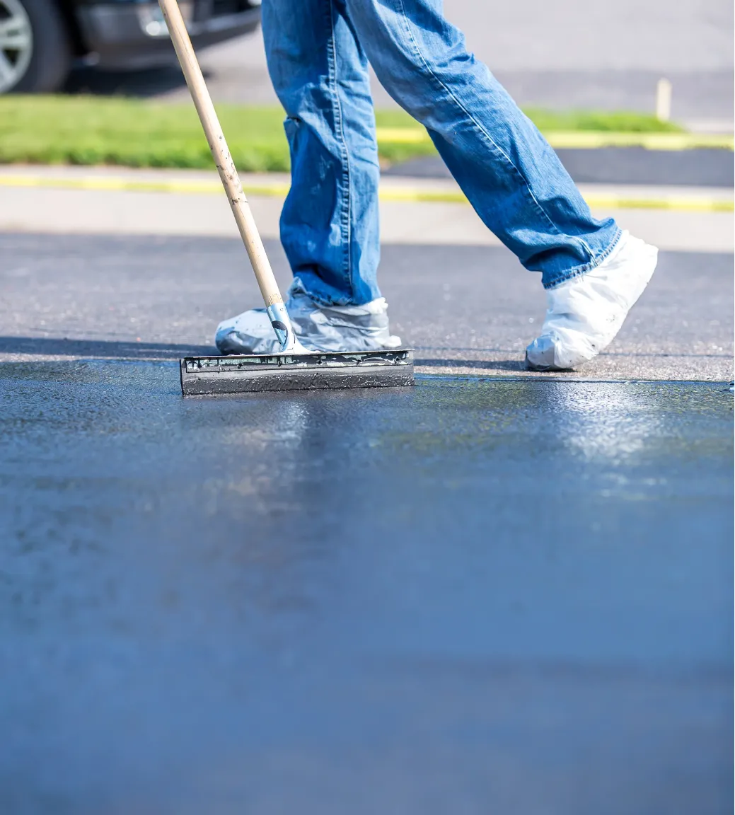 sealcoating-3 Person applying sealant to asphalt pavement with a squeegee, wearing jeans and shoe covers.