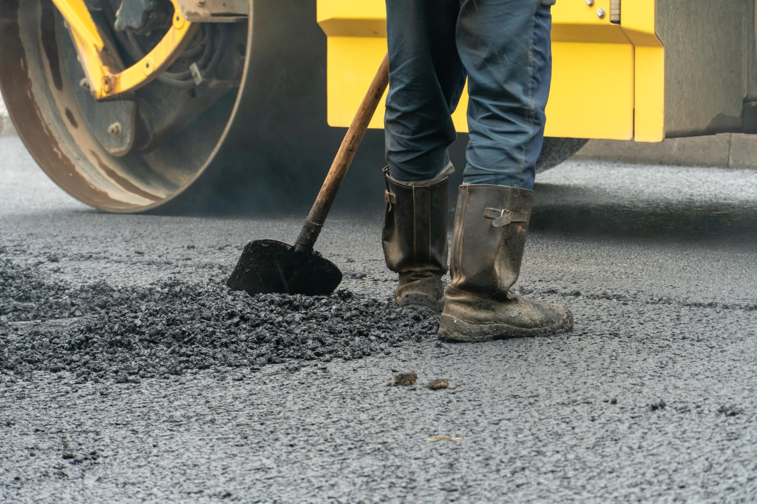 5440 Person in boots spreading asphalt on a road with a shovel near a yellow steamroller.